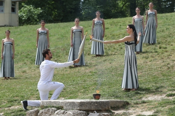 扮演最高女祭司的演員瑪麗·米娜（前右）點(diǎn)燃第一棒火炬手——希臘運(yùn)動(dòng)員、東京奧運(yùn)會(huì)男子賽艇項(xiàng)目金牌得主斯特凡諾斯·恩圖斯科斯手中的火炬。新華社記者 趙丁喆 攝